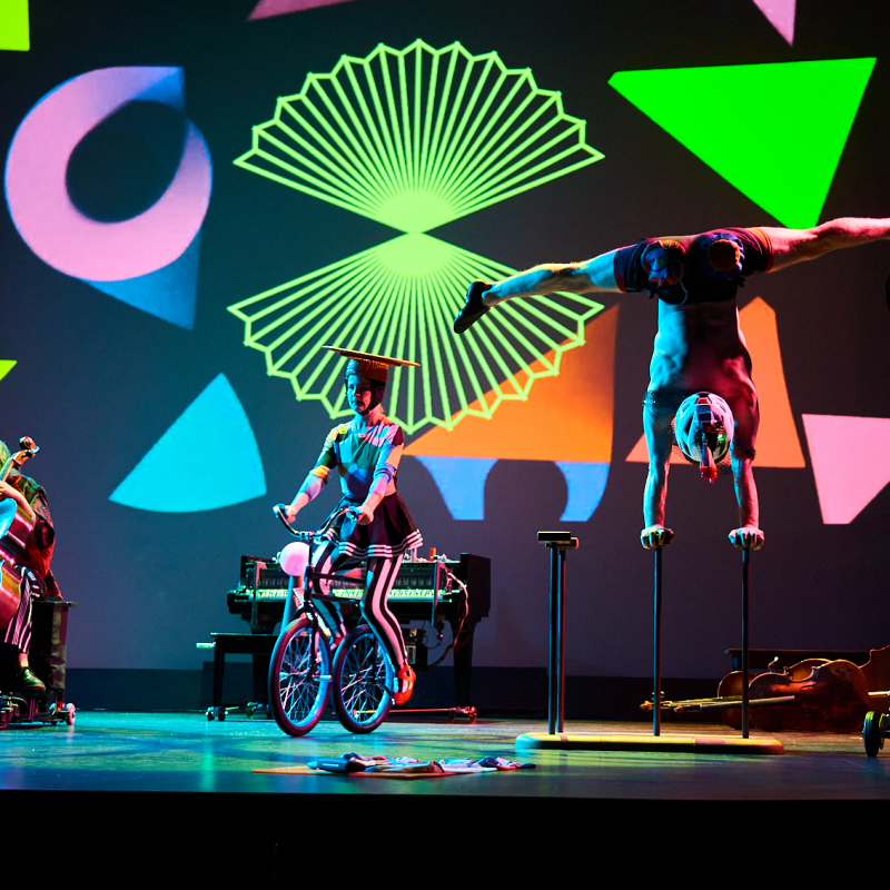 A cellist, a woman riding a bicylce, and a man doing an hand stand.